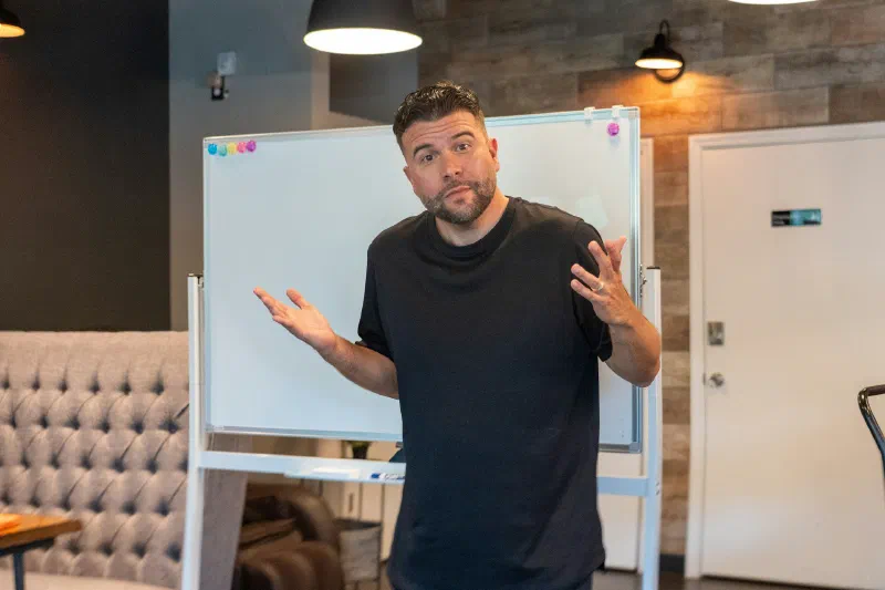 A man in a black T-shirt gestures with both hands while standing in front of a whiteboard in a modern office meeting room