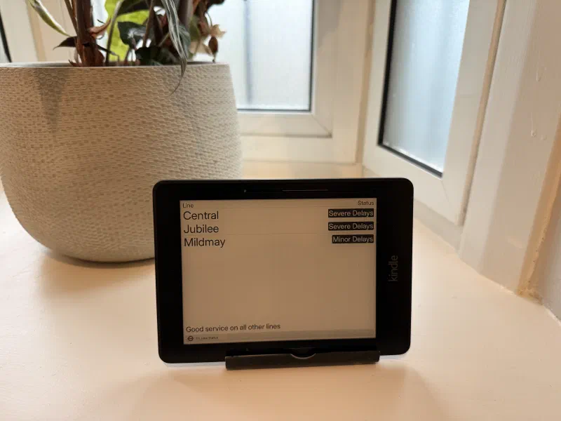 A Kindle Voyage on a windowsill displaying the TfL tube status dashboard. Central, Jubilee and Mildmay are all disrupted. A plant pot in the background.